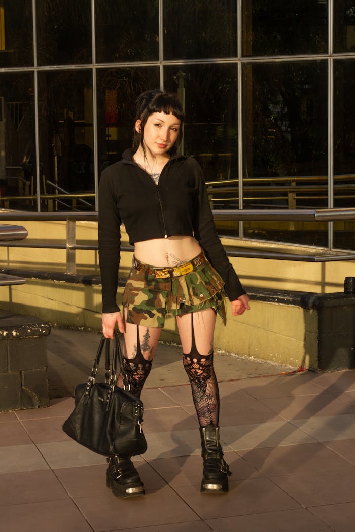 Young woman in chic street style with lace tights and camo skirt, posing in sunny urban setting.