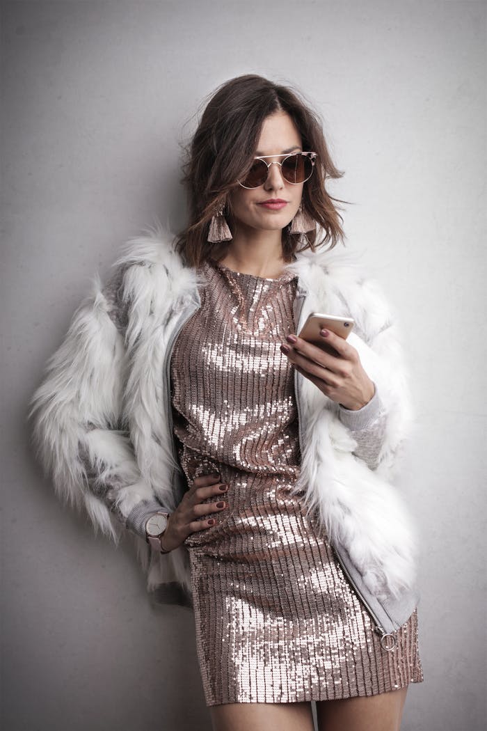 Stylish woman in sequined dress and fur coat texting on smartphone against a gray wall.